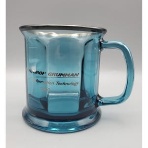 May include: A translucent blue glass mug with a sturdy handle and a wide base. The front surface features the printed text NORTHROP GRUMMAN Information Technology TASC in a dark font. The mug has a smooth, reflective finish and a cylindrical shape.