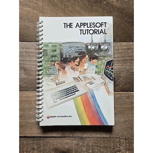 May include: A spiral-bound instructional manual titled The AppleSoft Tutorial. The cover features a vintage illustration of people using early Apple computer equipment, including a keyboard and monitor, with a rainbow stripe graphic at the bottom.