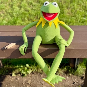 May include: A plush Kermit the Frog doll, seated on a wooden bench. The green frog has a yellow collar, red mouth, and white eyes. Two small wooden sticks are to the left of the frog. The background is blurred green grass.