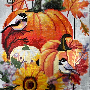 May include: A close-up of a fall-themed diamond painting featuring a large orange pumpkin with two small birds perched on it. The pumpkin is surrounded by sunflowers, white flowers, and fall leaves.