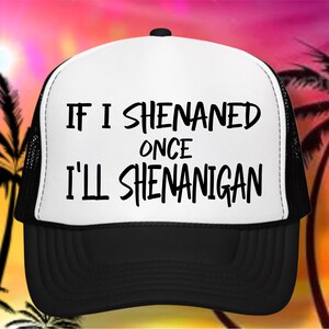 May include: A black and white trucker hat with the text "IF I SHENANED ONCE I'LL SHENANIGAN" in black. The hat has a white front panel and a black brim and mesh back. The background features a sunset with palm trees.