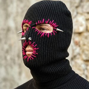 May include: A black knitted balaclava featuring bright pink embroidered stitching around the eye and mouth openings. The headwear is decorated with three silver-toned metal spikes positioned near the eye and nose areas of the knitted fabric.