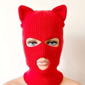 Pode incluir: Uma balaclava de malha vermelha brilhante com duas orelhas pontiagudas semelhantes às de um gato no topo. O acessório é construído com um padrão de malha canelada e inclui duas aberturas circulares para os olhos e uma abertura maior.