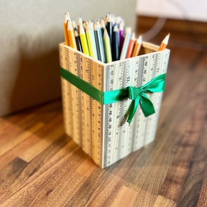 May include: A wooden pencil holder made from multiple rulers, decorated with a green ribbon bow. The holder is filled with coloured pencils.