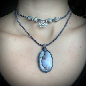 May include: A layered necklace with a grey cord and a silver lotus charm. The bottom layer features a large oval pendant with a white and grey agate stone.