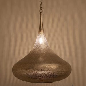 Pode incluir: Uma luminária pendente dourada com formato de gota. A tela de metal é perfurada com pequenos orifícios, permitindo que a luz filtre. A luminária pende de uma corrente e tem um topo pontiagudo. A marca "NUBARI LIGHTS" é visível.
