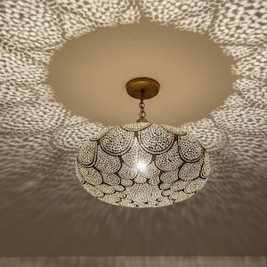 Puede incluir: Una lámpara colgante ornamentada con un diseño estampado. La luz proyecta intrincadas sombras en el techo, creando un efecto decorativo. La lámpara está suspendida de un soporte de techo dorado.