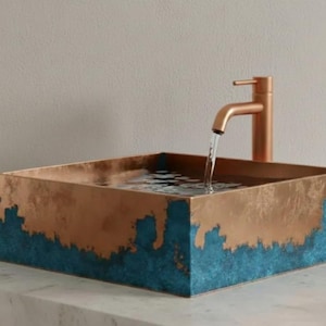 Könnte beinhalten: Ein quadratisches Waschbecken in Kupfer mit türkisfarbenem Rand. Ein Wasserhahn aus Kupfer befindet sich an der Seite, wobei Wasser in das Waschbecken fließt. Das Waschbecken ist mit Wasser gefüllt. Modernes Design.