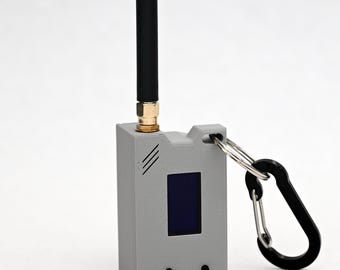 May include: A rectangular gray electronic device featuring a vertical black antenna, a small dark display screen, two circular buttons, and a black carabiner attached to a metal ring on the side. The casing has three diagonal ventilation slits.