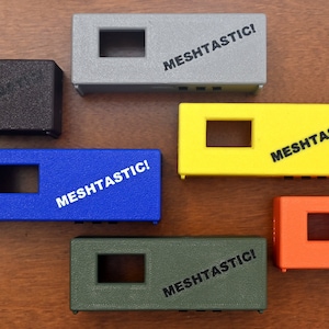 May include: Six small rectangular boxes in different colors, including black, gray, blue, yellow, orange, and green. Each box has a rectangular opening and the text "MESHTASTIC!" printed on the top.