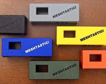 May include: Six small rectangular boxes in different colors, including black, gray, blue, yellow, orange, and green. Each box has a rectangular opening and the text "MESHTASTIC!" printed on the top.