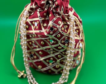 Puede incluir: Una bolsa burdeos con cordón, detalles dorados y un patrón geométrico. La bolsa está adornada con pequeños espejos y un hilo de cuentas. El cordón es dorado, sobre un fondo verde.