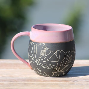May include: A ceramic mug with a pink interior and handle, and a black exterior. The black exterior features a white floral design. The mug is sitting on a wooden surface.
