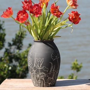 May include: A black ceramic vase with a floral design holds a bouquet of vibrant red tulips. The vase is cylindrical with a slightly flared top. The background features a body of water and green foliage.