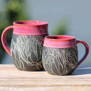 Puede incluir: Una taza de cerámica blanca con un asa negra y el texto "But First, Coffee" en negro, con una fuente de estilo manuscrito. La taza está sobre una superficie blanca.