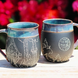 May include: Two ceramic mugs with a matte black finish and a glossy blue-green rim. The mugs feature white line art engravings of a forest scene with treehouse structures on one and a spherical space station above a forest on the other.