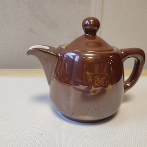 May include: A brown ceramic teapot with a lid and a gold design on the side. The teapot has a spout and a handle.