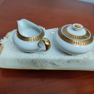 May include: A white porcelain creamer and sugar bowl set with gold trim. The set is on a white lace doily and a white tray with a gold rim.