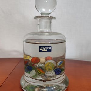 Könnte beinhalten: Eine klare Glasflasche mit einem runden Stopfen, gefüllt mit bunten Murmeln. Die Flasche hat ein Etikett mit der Aufschrift "LEONARDO".