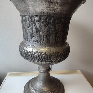 Könnte beinhalten: Eine silberne Metallvase mit einem verzierten Design mit Figuren und floralen Motiven. Die Vase hat eine breite Öffnung und einen hohen, schlanken Sockel.