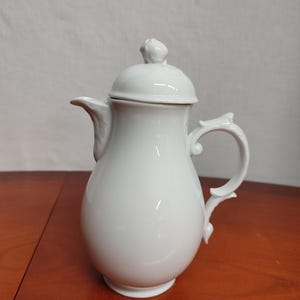 Può includere: Lattiera in porcellana bianca con coperchio e manico delicato. La lattiera ha un corpo arrotondato e un motivo floreale sul manico.