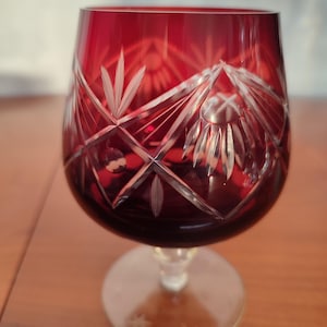 Può includere: Un bicchiere da brandy in cristallo rosso rubino con intricati disegni intagliati. La coppa del bicchiere è di un rosso intenso, che sfuma nel trasparente sullo stelo e sulla base. La base ha un diametro di circa 7,6 cm.