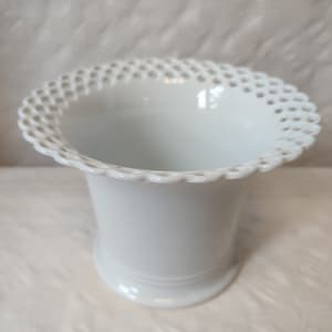 Può includere: Vaso in ceramica bianca con un bordo decorativo a pizzo. Il vaso ha una forma semplice e cilindrica.