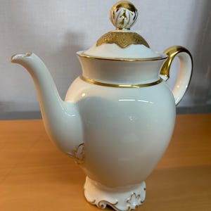 Puede incluir: Una tetera de porcelana blanca con adornos y detalles dorados. La tetera presenta un pico curvo, un cuerpo redondeado y una tapa con un pomo dorado decorativo. El asa y la base también tienen detalles dorados.