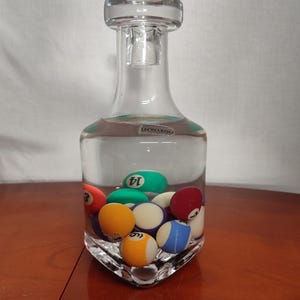 Könnte beinhalten: Eine klare Glaskaraffe mit quadratischem Boden und rundem Stopfen. Die Karaffe ist mit Wasser gefüllt und enthält mehrere bunte Billardkugeln.