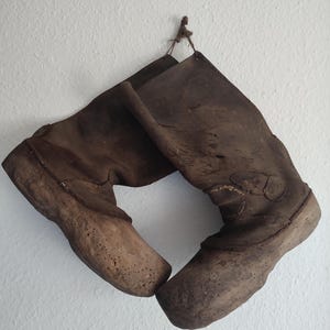 May include: A pair of brown leather boots with cork soles hanging on a white wall. The boots are worn and have a rustic look.