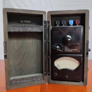 Könnte beinhalten: Eine Holzkiste mit einem schwarzen elektrischen Gerät darin. Das Gerät hat ein Zifferblatt, einen Knopf und drei schwarze Knöpfe mit roten, blauen und schwarzen Kappen. Die Kiste ist mit "Ln 17024" beschriftet.