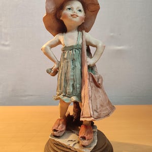 Könnte beinhalten: Eine Keramikfigur eines Mädchens, das einen braunen Hut, ein blau-weißes Kleid und rote Schuhe trägt. Sie hält eine braune Tasche in ihrer rechten Hand und hat einen verspielten Gesichtsausdruck.