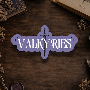 May include: A light purple sticker featuring the white text VALKYRIES with a sword graphic replacing the letter I, surrounded by small star accents. The sticker has a die-cut shape and is centered on a dark wooden surface with various vintage items.