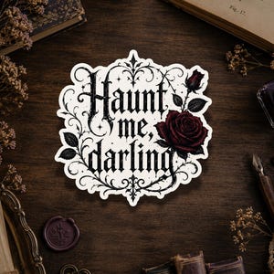 Peut inclure: Un autocollant découpé présentant le texte Haunt me, darling dans une police gothique noire, entouré de volutes en filigrane noir complexe et d'une rose rouge foncé avec des feuilles vertes, le tout sur un fond blanc à bord festonné.
