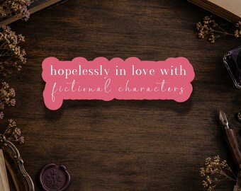 Könnte beinhalten: Ein rosafarbener, konturgestanzter Aufkleber mit dem weißen Text "hopelessly in love with fictional characters" in einer Mischung aus serifenloser Schrift und Schreibschrift, umgeben von alten Büchern, Trockenblumen, einem Metallschlüssel, einem Wachssiegel und einem Füllfederhalter.