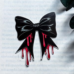 May include: A black bow sticker featuring a graphic design of bright red liquid dripping from the bottom edges of the ribbon tails. The bow has a central knot and wide, flared loops with white outlines, presented against a background of printed text.