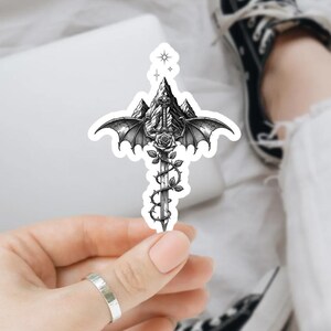 May include: A die-cut sticker featuring a black and white illustration of a sword entwined with a rose vine, positioned in front of a mountain range with large bat-like wings extending from the sides and a small four-pointed star at the top.