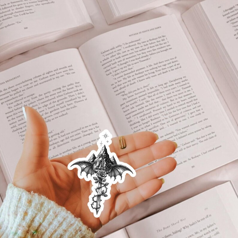 May include: A black and white sticker featuring a sword entwined with a rose, positioned in front of a mountain range with large bat-like wings extending from the sides and a small star symbol at the top, all rendered in a detailed line art style.
