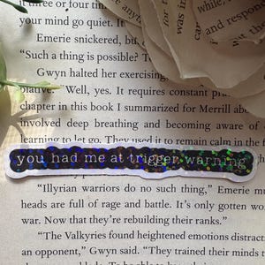 以下が含まれることがあります： 青いステッカーで「you had me at trigger warning」というテキストが強調表示された本のページのクローズアップ。ページのテキストは、イリュリアの戦士とその訓練について説明しています。