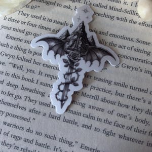May include: A black and white sticker featuring a central sword design with large bat wings extending from the hilt. The blade is wrapped in thorny vines and a rose, with a mountain range depicted behind the wings and a small star icon at the top.