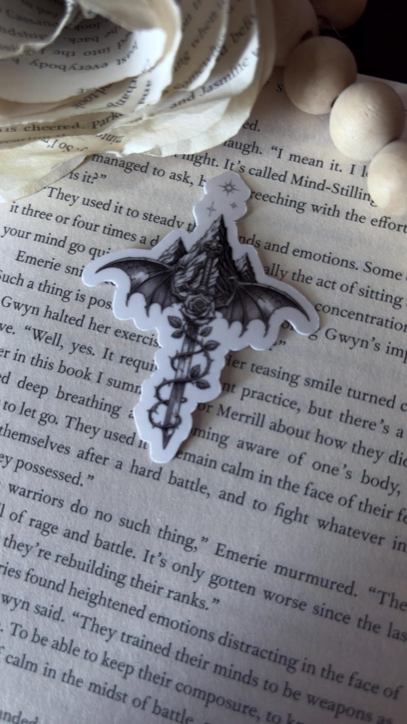 May include: A black and white sticker featuring a central sword design with large bat wings extending from the hilt. The blade is wrapped in thorny vines and a rose, with a mountain range depicted behind the wings and a small star icon at the top.