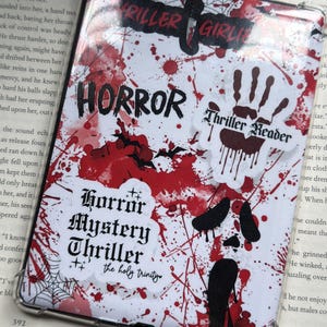 May include: A clear tablet case with a white background covered in red splatter and horror-themed stickers. The stickers include the words "HORROR," "Thriller Girl," and "Thriller Reader." A book is visible in the background.