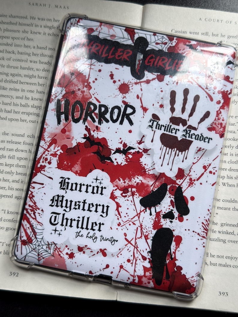 May include: A clear tablet case with a white background covered in red splatter and horror-themed stickers. The stickers include the words "HORROR," "Thriller Girl," and "Thriller Reader." A book is visible in the background.