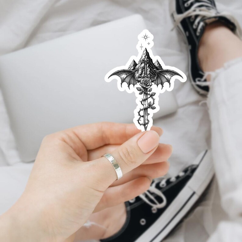 May include: A die-cut sticker featuring a black and white illustration of a sword entwined with a rose vine, positioned in front of a mountain range with large bat-like wings extending from the sides and a small four-pointed star at the top.
