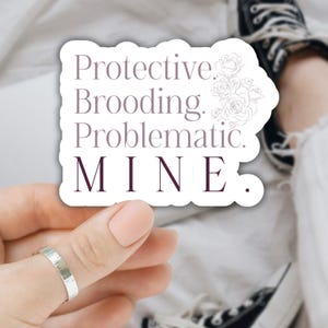 May include: A white sticker with the words "Protective. Brooding. Problematic. MINE." in a dark purple font. A floral design is in the upper right corner. A silver ring is on a finger.