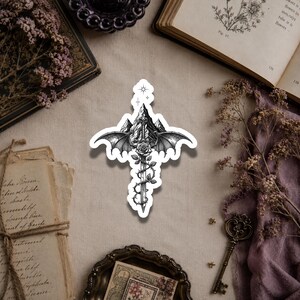 May include: A die-cut sticker featuring a black and white ink illustration of a sword centered in front of a mountain range. The sword is adorned with a rose and thorny vines, flanked by two large bat wings, and topped with a small four-pointed star.