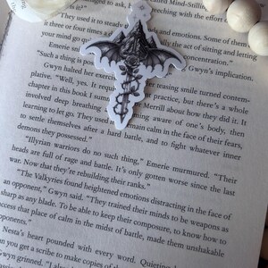 May include: A die-cut sticker featuring a black and white illustration of a sword with bat wings, a rose wrapped around the blade, and a mountain peak at the hilt. The sticker is placed on an open book page with printed text, near a paper flower.