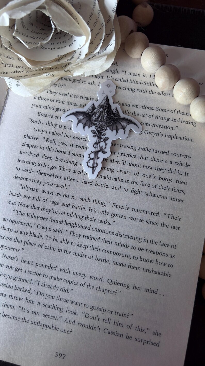 May include: A die-cut sticker featuring a black and white illustration of a sword with bat wings, a rose wrapped around the blade, and a mountain peak at the hilt. The sticker is placed on an open book page with printed text, near a paper flower.