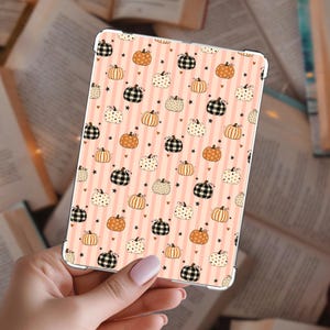 Op de afbeelding: Een rechthoekige tablethoes met een herhalend patroon van oranje, witte en zwarte geruite pompoenen, afgewisseld met kleine zwarte sterretjes en hartjes op een achtergrond van verticale lichtperzik en witte strepen.