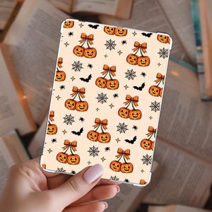 Op de afbeelding: Een rechthoekige tablethoes met een lichtcrème achtergrond voorzien van een herhalend patroon van oranje pompoenen in de vorm van kersen met oranje strikken, zwarte spinnenwebben, kleine zwarte spinnen, zwarte vleermuizen en kleine vierpuntige sterren.